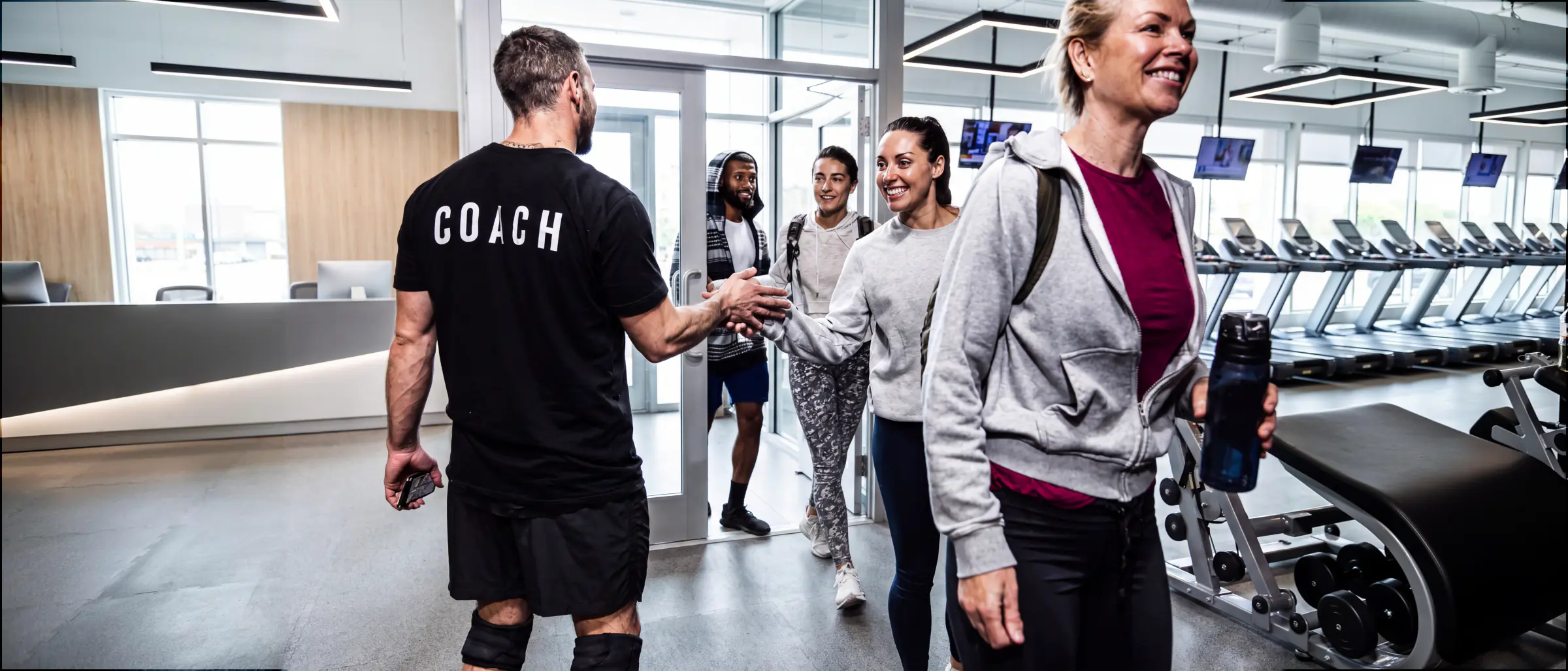 A coach shaking hands with an arriving member at the entrance of a bright commercial gym, more members walking in behind them past a row of treadmills